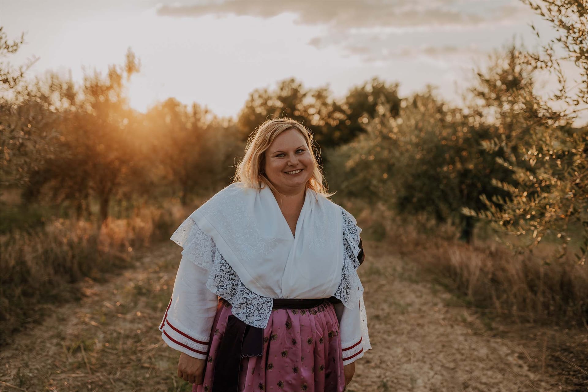 Ženska v narodni noši se smehlja na poti med olivami ob sončnem zahodu.