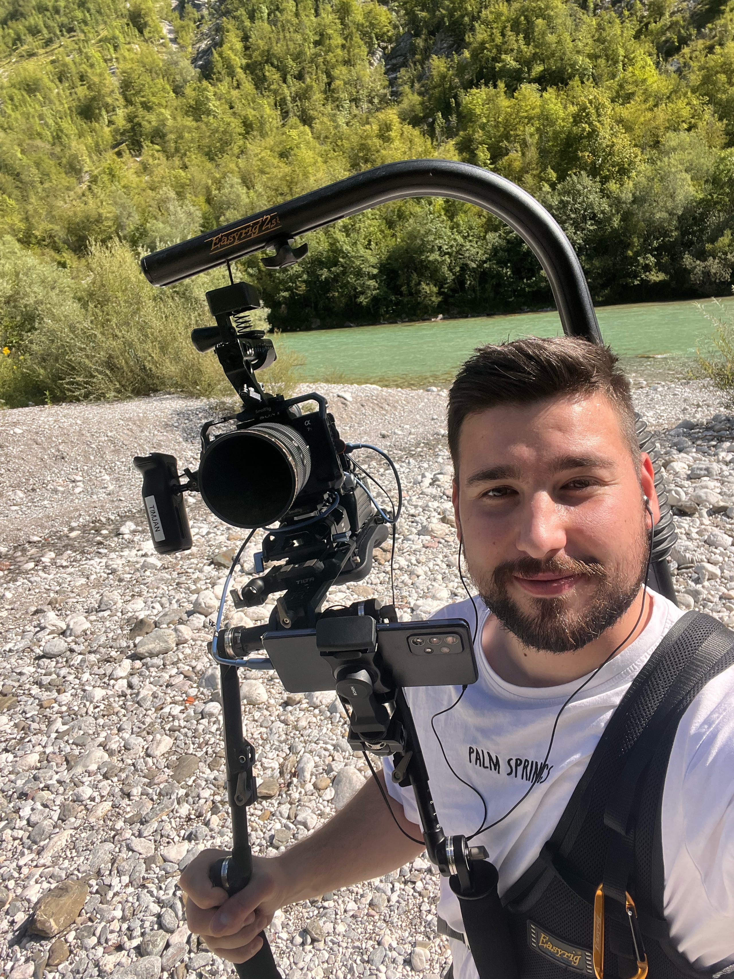 Moški s kamero na gimbal stabilizatorju stoji ob obali reke.