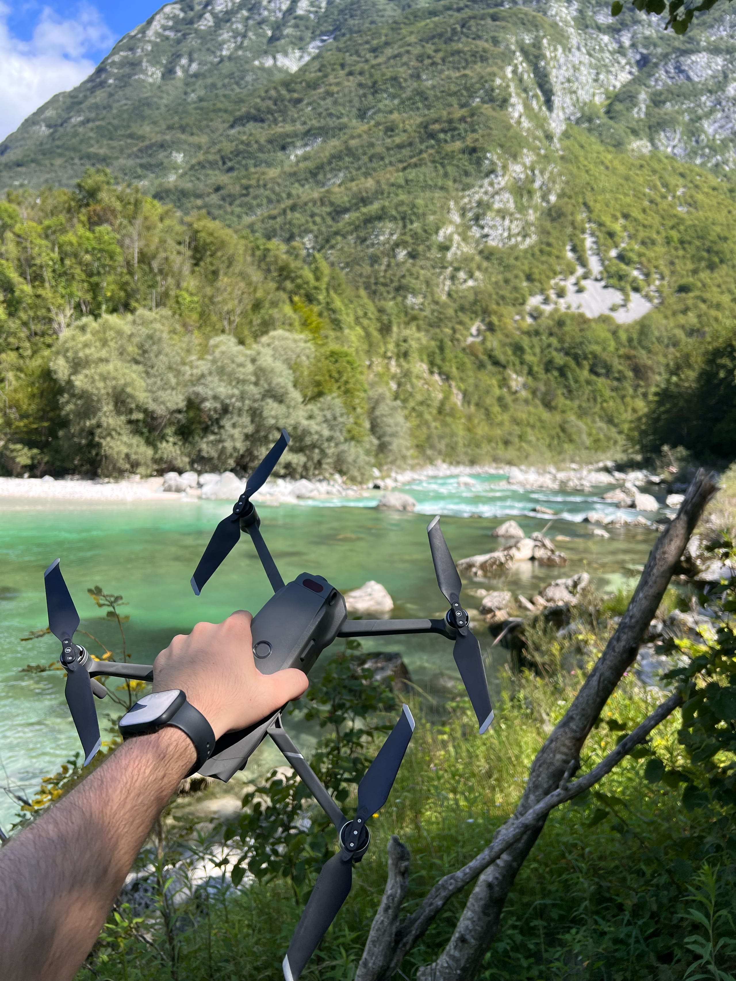 Roka drži dron nad turkizno reko med gorami in gozdom.
