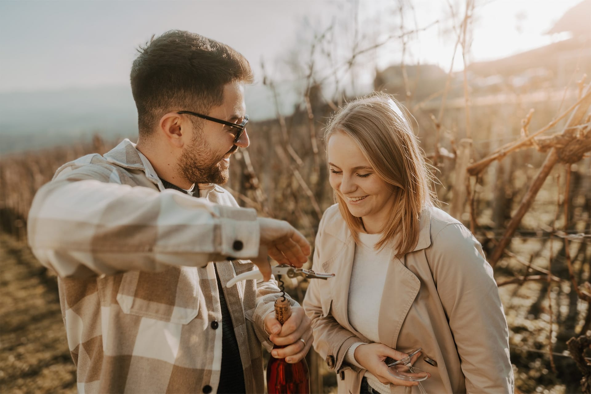 Moški in ženska med odpiranjem steklenice vina v vinogradu ob sončnem zahodu.