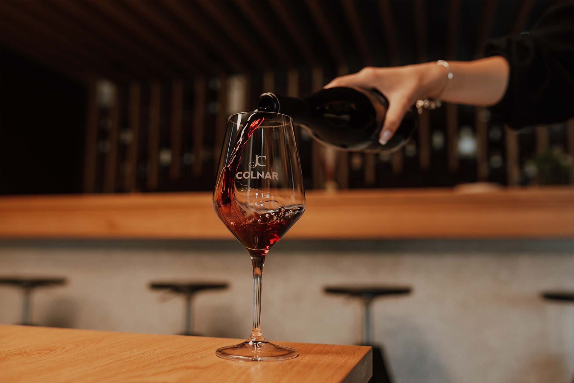 Roka toči rdeče vino v kozarec z napisom Colnar na leseni mizi.