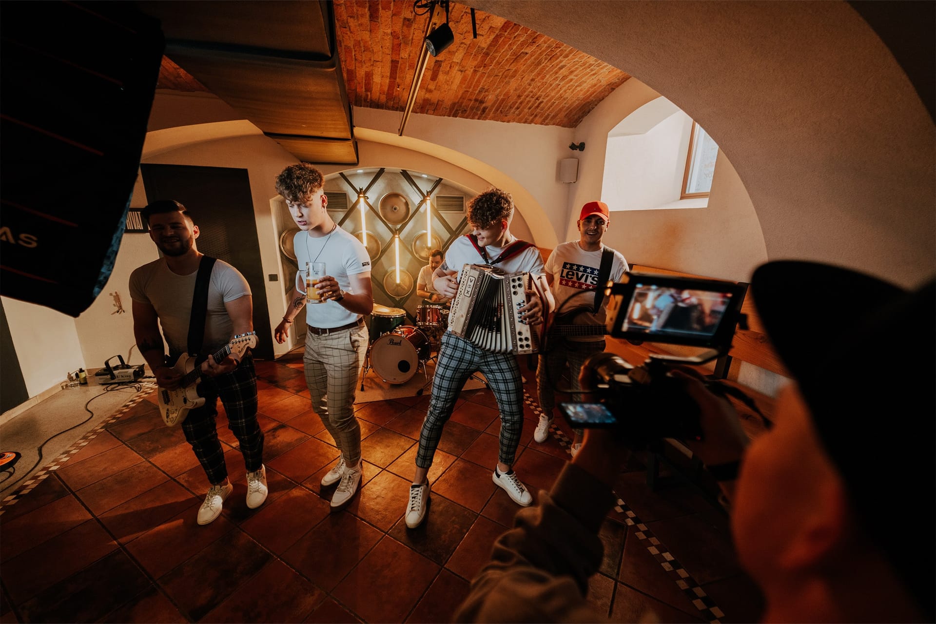 Glasbeniki nastopajo v obokanem prostoru med snemanjem videospota, ekipa opazuje monitor.