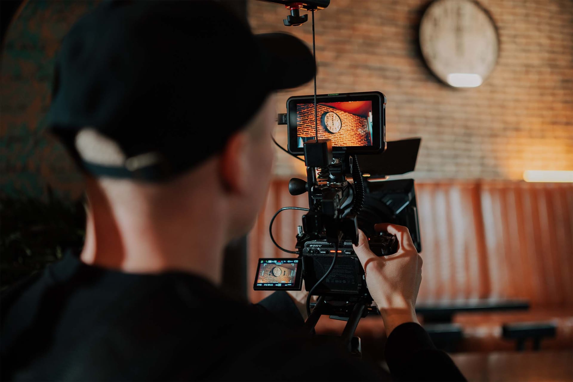 Snemalec s kapo snema z videokamero v prostoru z opečnato steno.