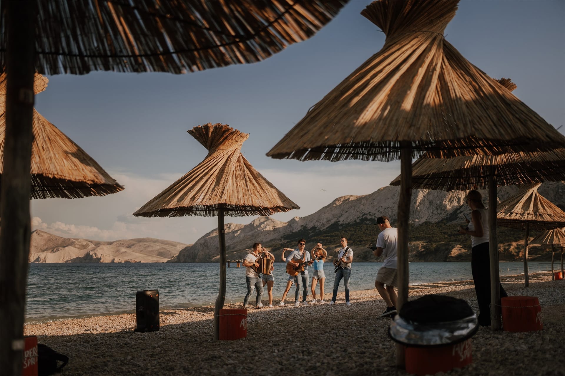 Glasbeniki igrajo na plaži pod slamnatimi senčniki ob morju in gorah.