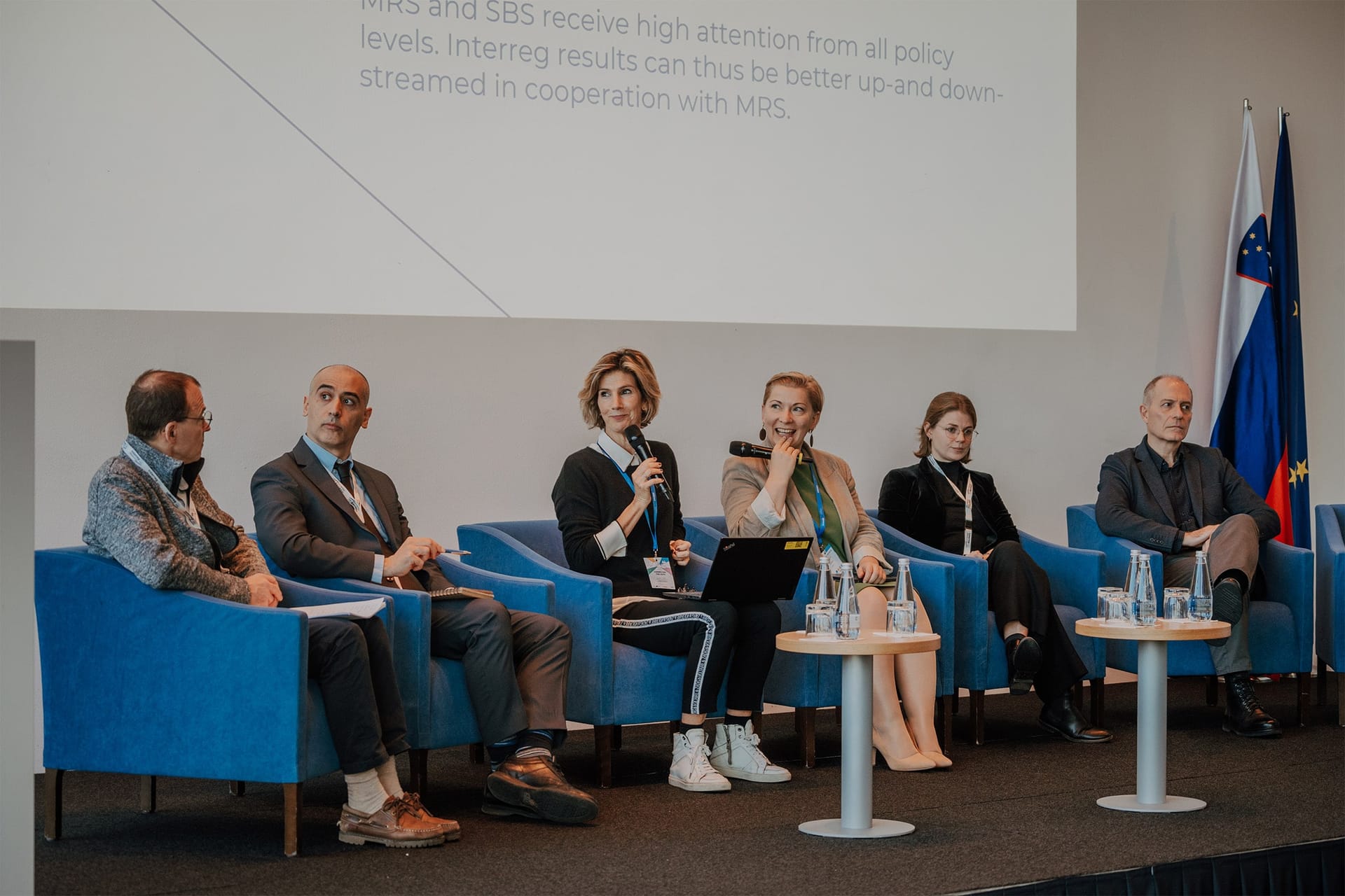 Šest govorcev na panelni razpravi sedi na modrih stolih pred projekcijskim platnom.