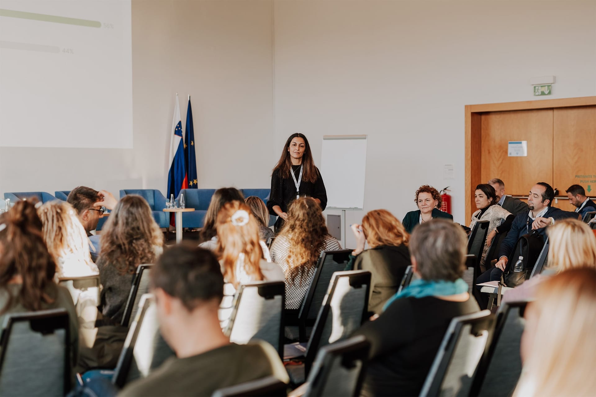 Predavateljica govori pred publiko v konferenčni dvorani z evropsko in slovensko zastavo.