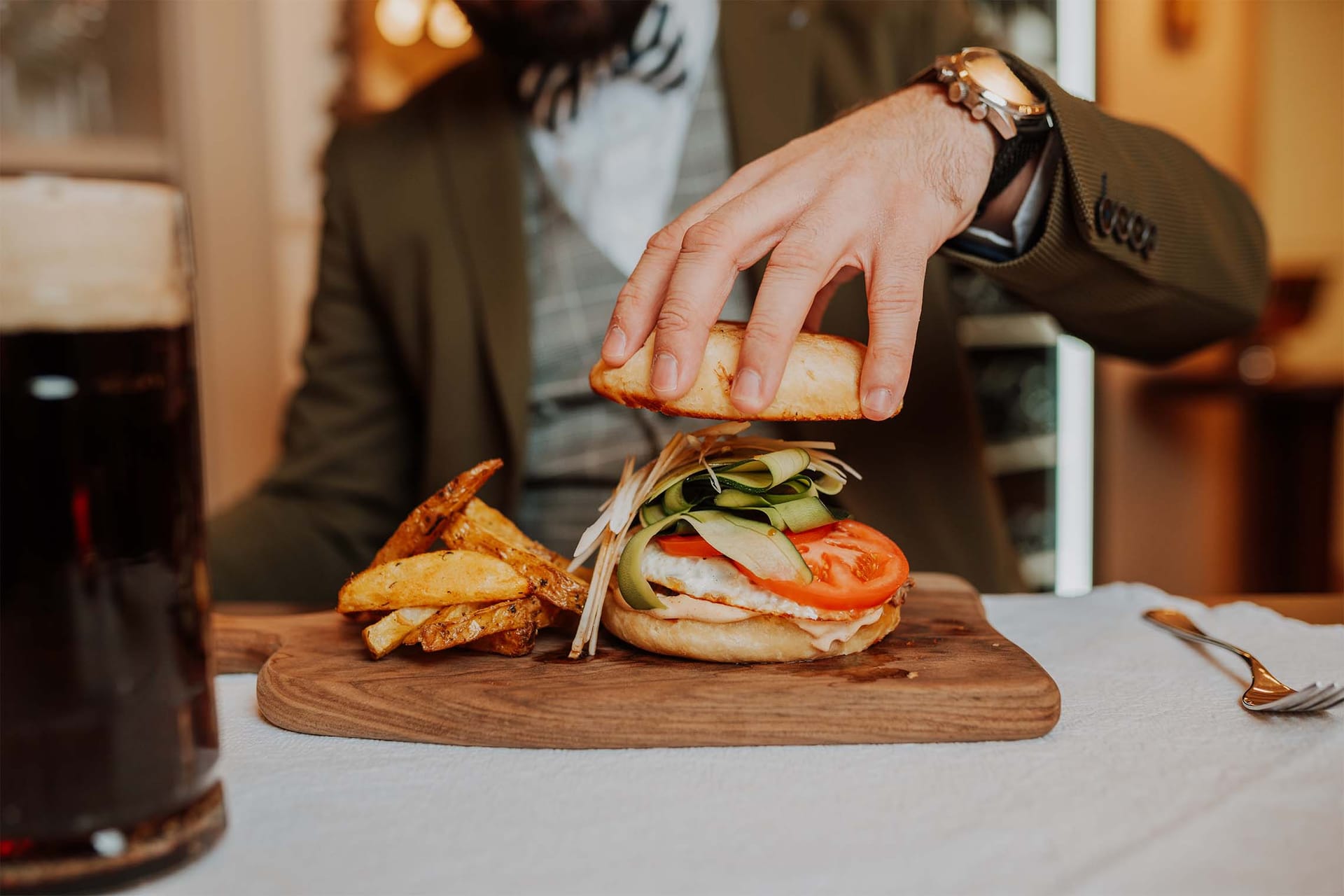 Moški sestavlja burger s krompirjem in pivom na leseni deski.