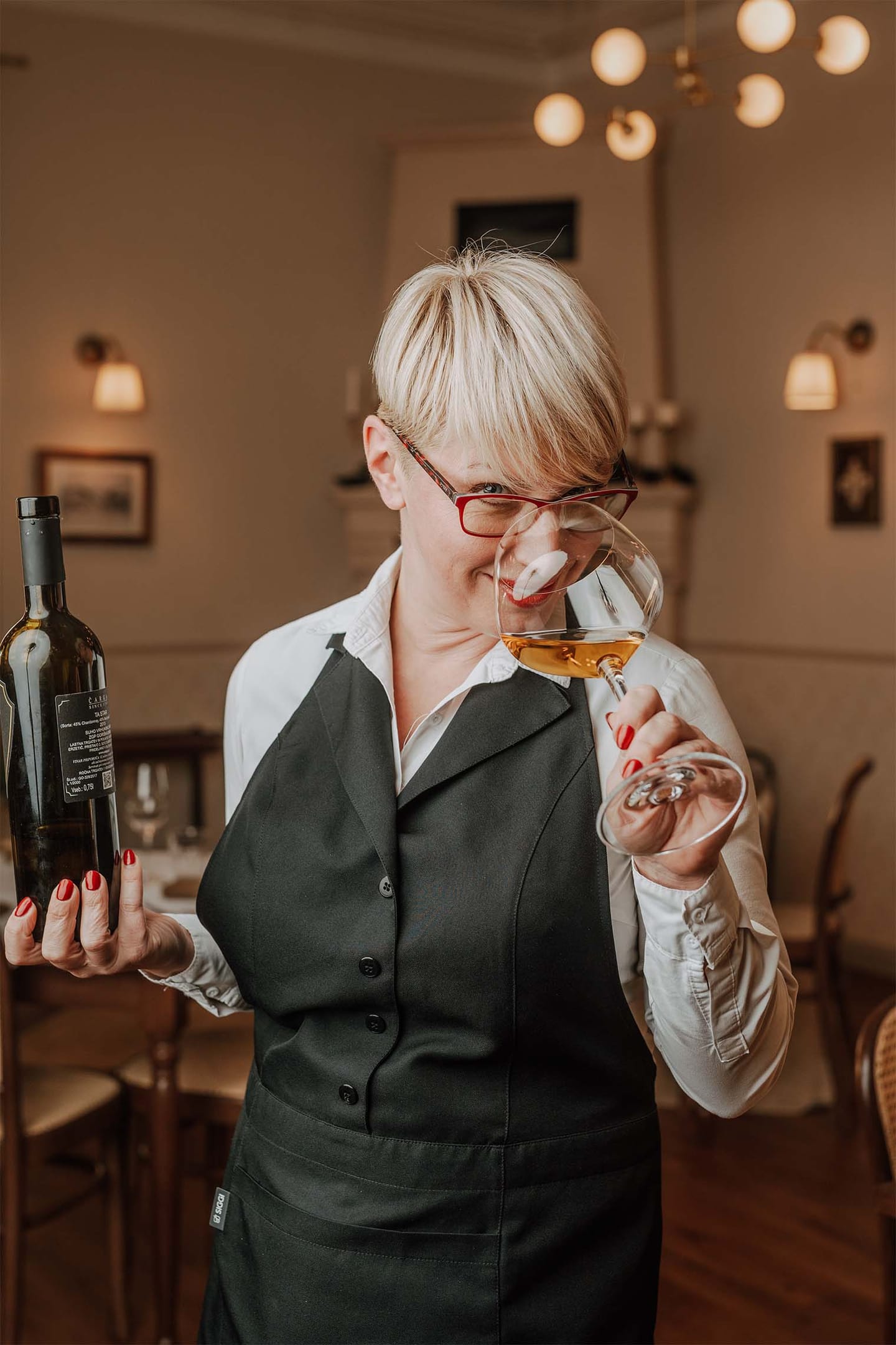 Natakarica v predpasniku degustira vino iz kozarca in drži steklenico v elegantni restavraciji.