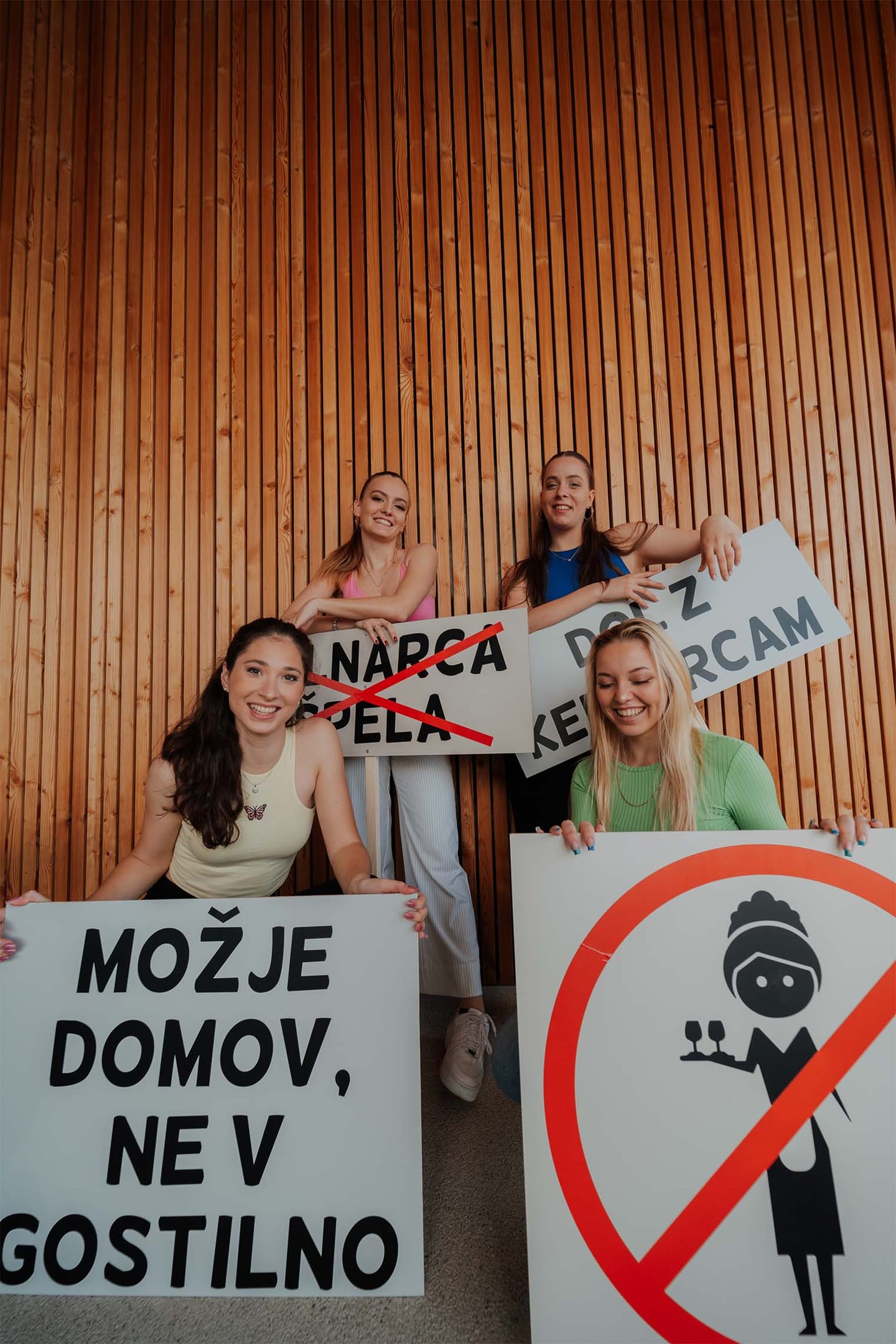 Štiri nasmejane ženske pred leseno steno držijo šaljive napise s protestnimi gesli.