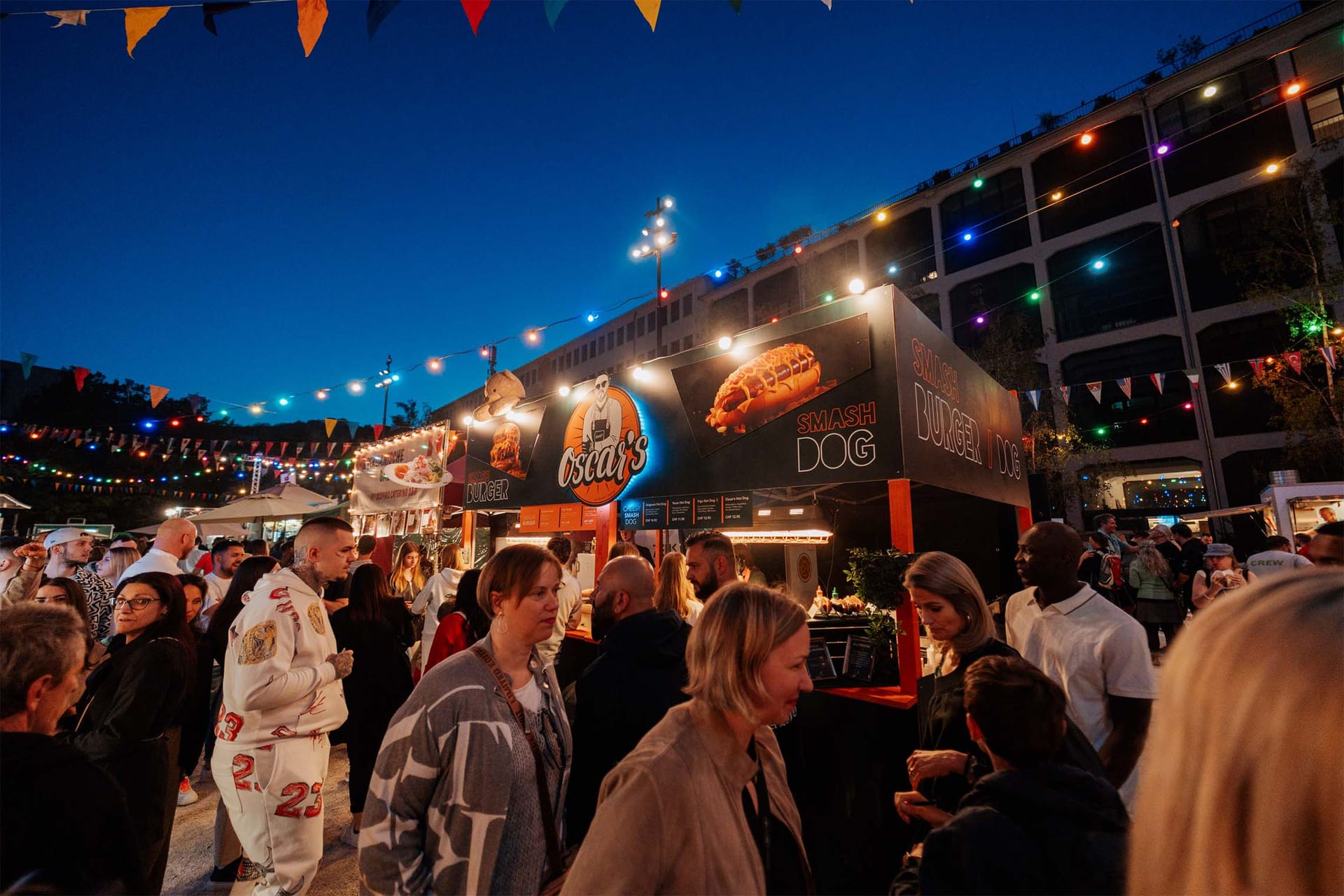 Množica ljudi na večernem festivalu hrane pod girlandami lučk in zastavicami.
