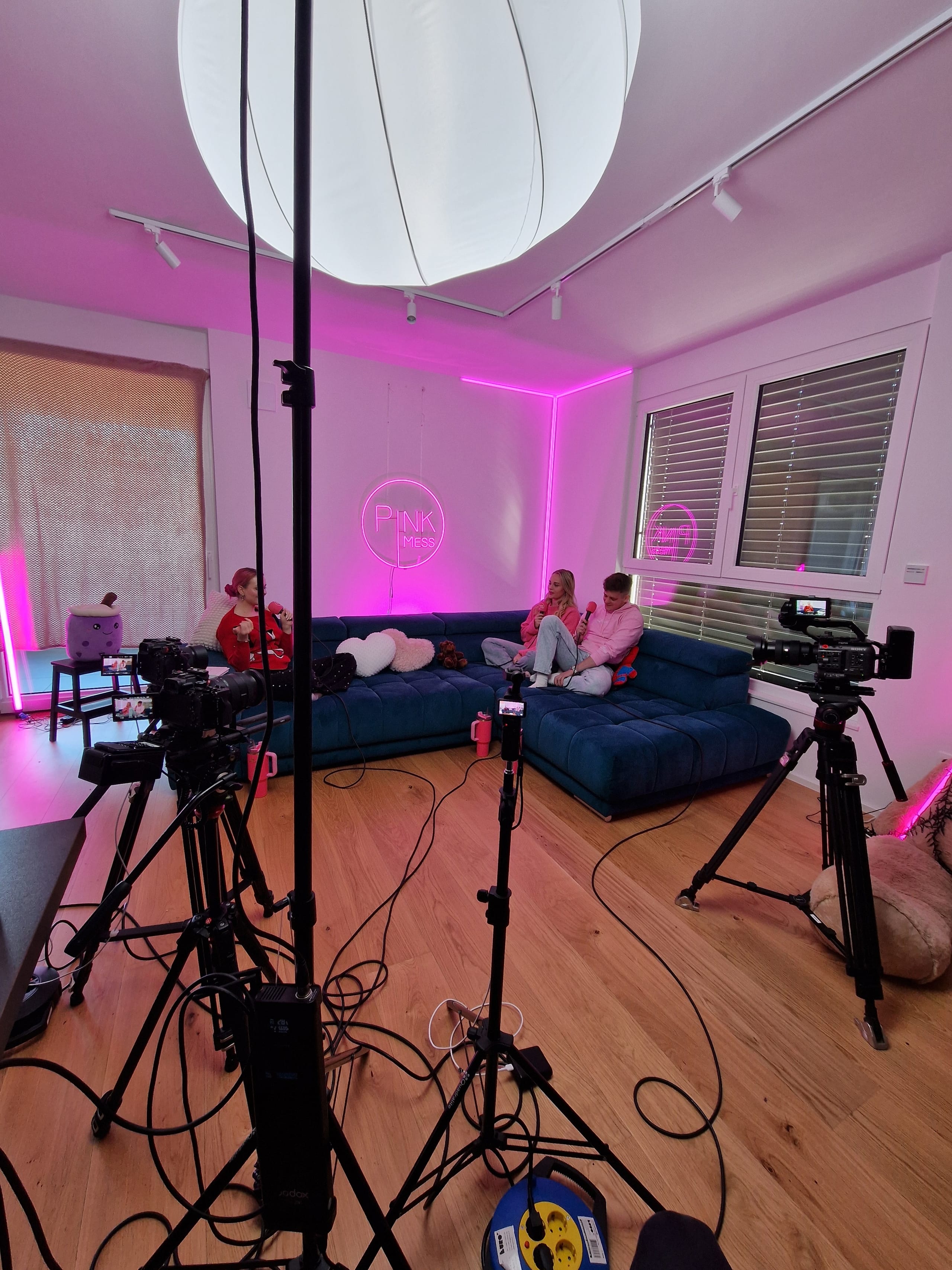Snemalni studio z roza osvetlitvijo, kamerami, mikrofoni in modrim kavčem.