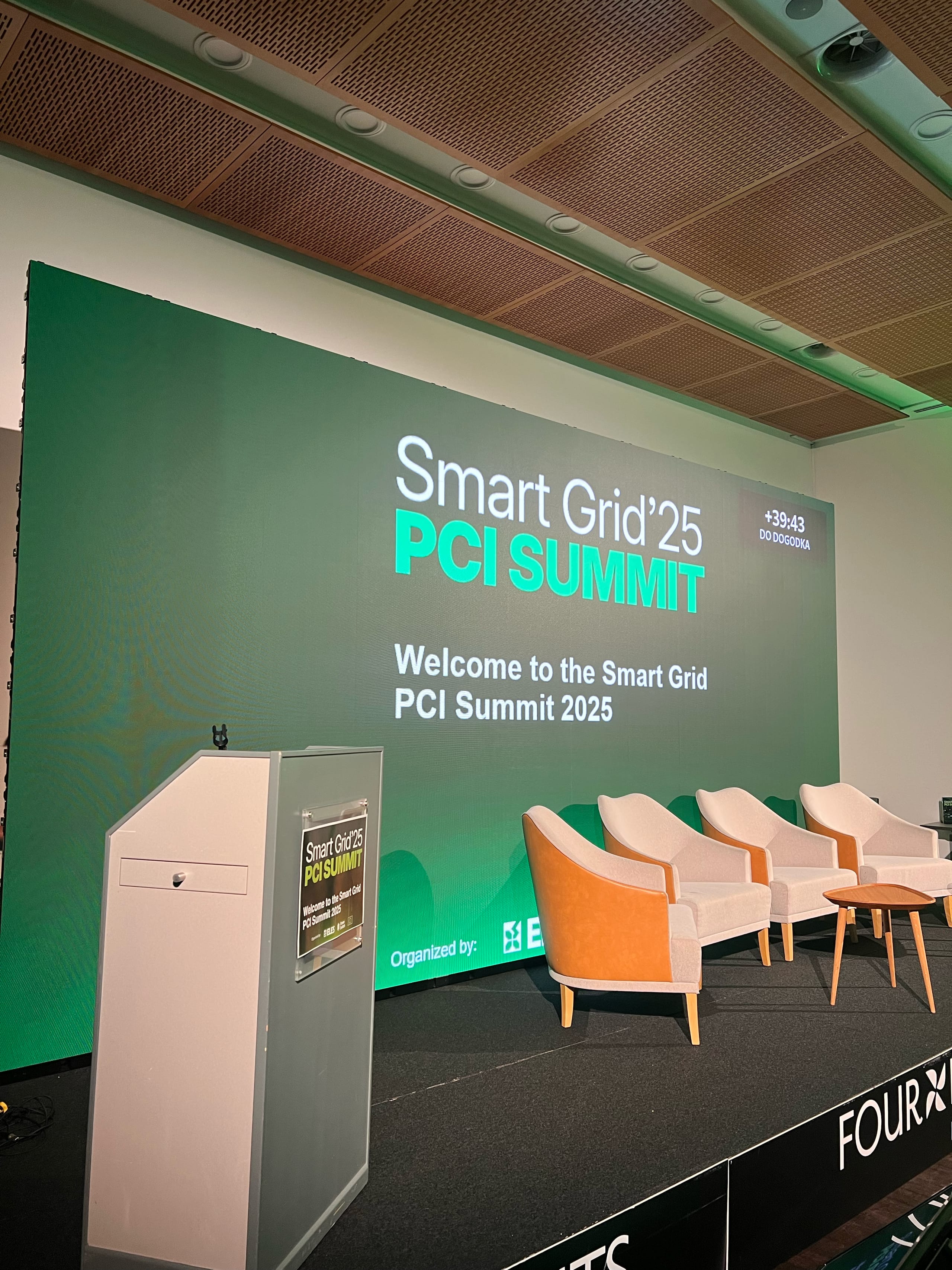 Oder z LED zaslonom in napisom Smart Grid PCI Summit, oranžni stoli v ozadju.