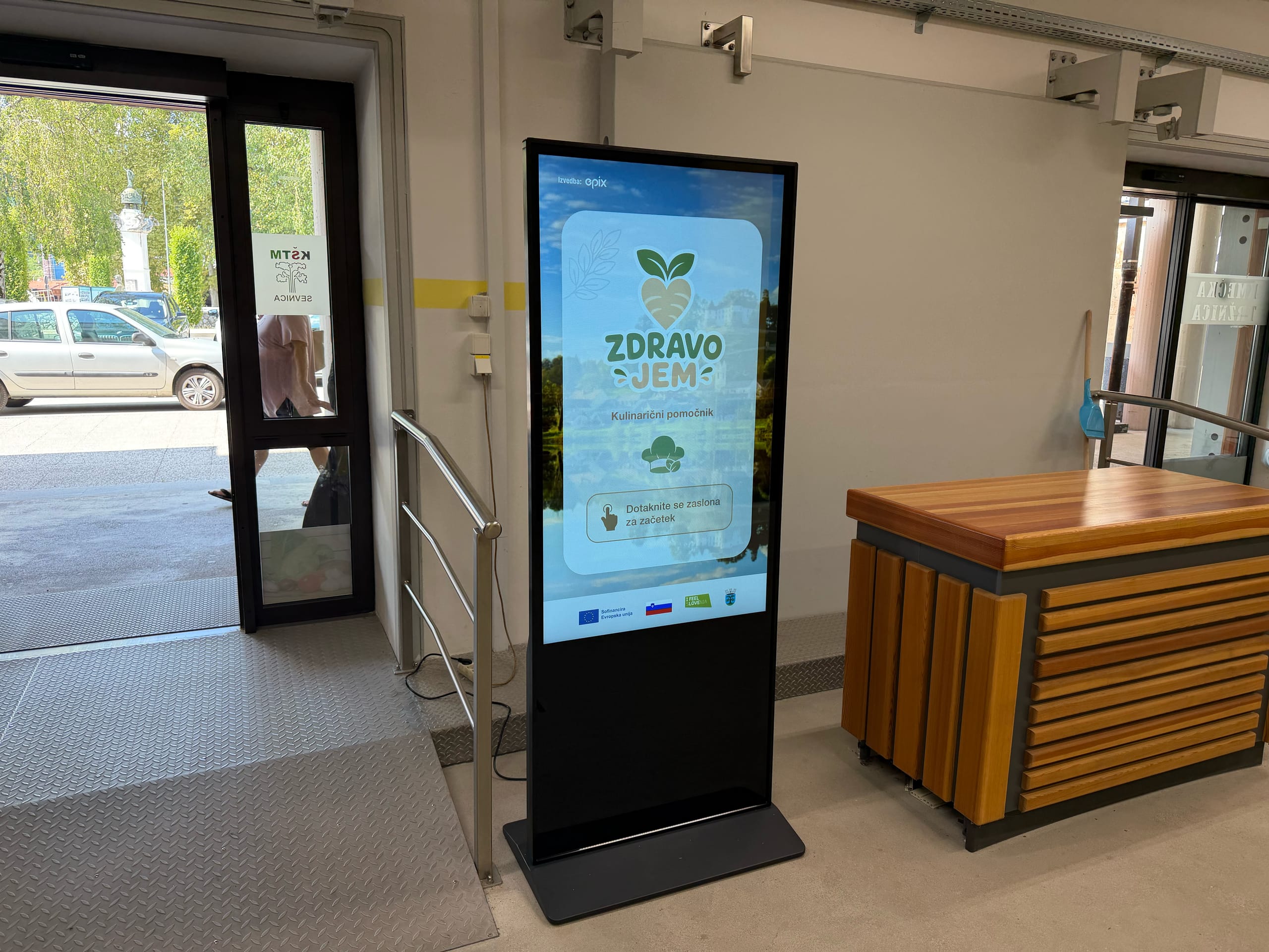 Digitalni zaslon z napisom Zdravo jem stoji ob vhodu v stavbo.
